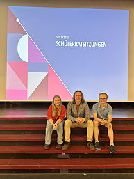 Das Foto zeigt Saskia Lübbers, Leon Wenke und Hauke Wasse auf den Stufen zur Bühne in der Aula des TGG bei der Schülerratssitzung am 22.05.2024.